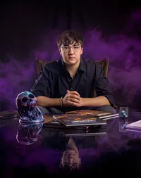 Professional photography by Nature's Child Photography: Senior poses in a Mandarin studio with swirling purple smoke, hands folded at a reflective table beside books and a decorative skull in a dramatic senior portrait session.