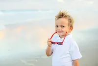 Ponte Vedra portrait session capturing Young boy with wind-tousled blond hair holding red sunglasses and flashing a confident grin during a beach child portrait session.