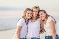 Jacksonville, FL portrait session captured at sunrise on Micklers Beach, a mother pulls her two teenage daughters close, their heads touching and smiles easy during a family portrait session.