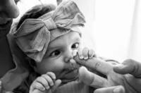 Professional photography by Nature's Child Photography: In Mandarin, a newborn with a large bow headband stares wide-eyed while clutching a parent’s finger, her tiny fingers wrapped tight during a lifestyle newborn.