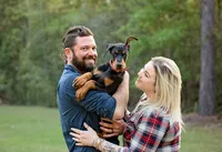 Professional photography by Nature's Child Photography: Middleburg, pet featuring a Doberman Pinscher puppy held between its new parents, the young dog facing forward with alert eyes and oversized paws.