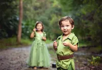 Jacksonville, FL portrait session capturing Smiling young boy in a green storybook-inspired outfit stands in the foreground as his sister in a flowing fairy dress holds a wand behind him during a fairy adventure signature session in Palatka.