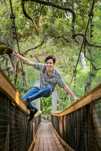 Jacksonville, FL portrait session capturing High school senior boy in Palatka, poses mid-balance on a tree-lined swinging bridge, blending movement and personality in a natural outdoor senior portrait session.