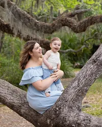 Professional photography by Nature's Child Photography: Mom and Me family featuring a mother perched in a wide tree branch, cradling her happy baby as soft green foliage fills the background.