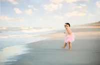 Baby girl in a pink tutu standing barefoot at the water’s edge, looking surprised as waves roll in during a beach child photography session- A portrait experience by Nature's Child Photography.