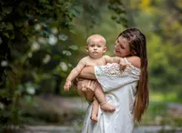 A mother gently lifts her baby boy against a backdrop of lush greenery and flowing water in Palatka, a sweet lipstick kiss pressed onto his forehead during their outdoor family session. - A portrait experience by Nature's Child Photography.