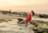 Palm Coast, photo of a senior high school graduate seated on seaside rocks at golden hour, her vibrant red dress contrasting against the soft ocean horizon. (Photographed by Nature's Child Photography)