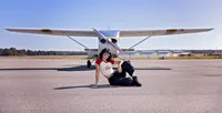 High school senior seated on the runway, framed by a small airplane behind her, captured in an editorial-style aviation senior photo session. - A portrait experience by Nature's Child Photography.