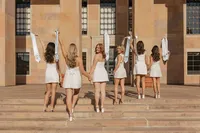 A group of girls in dressed in white stand in front of Norlin library & walk hand-in-hand up the steps as they wave their stoles for Boulder graduation photos.