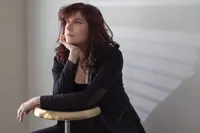 Woman with dark red curls in a black blazer seated against a white slatted wall, resting on a brass stool.Woman in a gold sequined dress seated on a geometric block, dark studio background scattered with warm points of light.