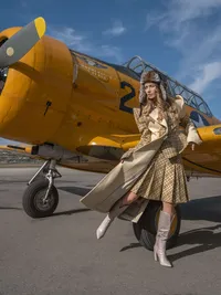 Model in layered gold and beige fashion standing beside a yellow vintage training aircraft.
