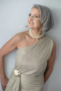 Woman with silver hair in a one-shoulder draped gold gown and pearl choker, leaning against a pale wall.