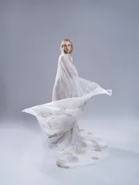 Model in a sheer white gown with the train fanned across the floor of a pale studio.