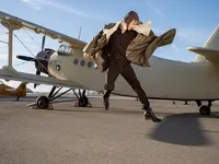 Person in a brown coat caught mid-movement beside a white biplane on an airfield tarmac.