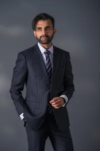 Man in a navy pinstripe suit and tie standing with hands in his pockets against a dark, moody background.Model in an embellished strapless gown with feather detailing, one arm raised, against a pale studio backdrop.