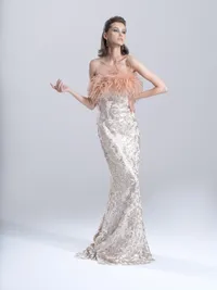 Model in an embellished strapless gown with feather detailing, one arm raised, against a pale studio backdrop.