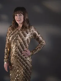Woman in a gold sequinned long-sleeve dress with one hand on her hip, dark background scattered with warm points of light.