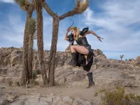 Model in dark clothing and boots posed on desert rock among Joshua trees under a bright blue sky.