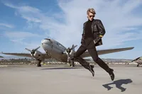 A person wearing a black leather jacket, black pants, and sunglasses is mid-jump in front of a vintage propeller airplane—captured in the bold style of a cinematic portrait photographer Washington DC, under blue skies with wispy clouds.