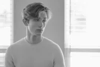 Black and white portrait of a young man in a light sweater standing beside a bright window.
