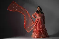 Woman in a red and gold embroidered lehenga, dupatta sweeping out to one side, one hand on her hip, against a grey studio backdrop.
