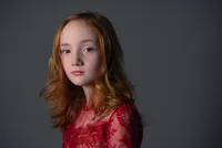 Portrait of a young girl with long red hair in a red lace dress against a dark grey backdrop.