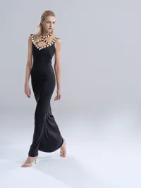 Model in a long black column gown with an embellished neckline, walking barefoot across a pale studio floor.