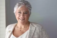 Smiling woman with short silver hair in a cream tweed jacket and gold necklace, photographed in bright natural light.Person in a black leather jacket and sunglasses caught mid-jump on an airfield tarmac, propeller aircraft behind.