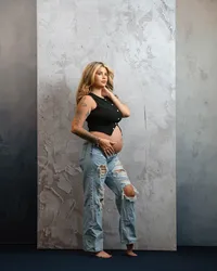 mom-to-be touching her hair and holding her bare belly, wearing a black top and ripped jeans.