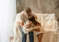 Father supporting his older daughter as she kisses her newborn sibling, surrounded by cream blankets and neutral colors.