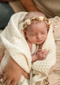 relaxed and peaceful newborn wearing a floral headband, nestled in her mother’s arms in a cream blanket with soft knitted textures.