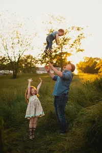 Father tossing his toddler into the air as his daughter cheers in tall grass during a playful golden hour family session.