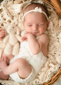 Smiling unposed newborn in a cream knitted outfit and white floral headband, resting in a wicker basket beside a soft toy.