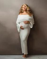 mom holding her belly in a fitted cream gown with draped shoulders during softly lit studio maternity portraits.