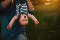 Toddler grinning upside down in his father’s arms during a playful outdoor family photoshoot in golden hour light.