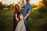 Smiling girl holding her parents’ hands while her father carries her baby brother in field at sunset