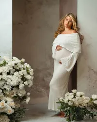 blonde mom in fitted cream gown touching belly standing near white romantic floral arrangements