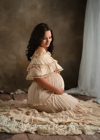 Pregnant woman kneeling in a cream ruffled gown, smiling down at her belly beside a softly lit studio window.