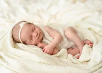 Sleeping newborn curled on her side in a cream knitted outfit and floral headband, surrounded by soft neutral blankets.