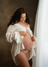 Pregnant woman smiling down at her belly in an open white shirt during a relaxed studio maternity session.