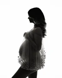Dramatic black-and-white studio maternity silhouette of a pregnant woman wearing a sheer embellished gown.