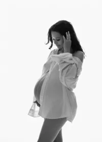 Black-and-white maternity portrait of a pregnant woman touching her hair in an open white shirt against a bright background.
