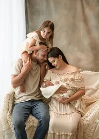 Parents cuddling their newborn while their older daughter sits on her father’s shoulders during a relaxed studio session.