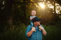 Smiling toddler riding on his father’s shoulders beneath sunlit trees during a relaxed outdoor family session.