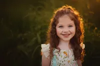 Close-up of a smiling girl in a floral dress, with golden hour sunlight illuminating her curls against a grassy background.