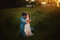 Father kneeling to hug his daughter in a flowing pink dress during a relaxed sunset family photoshoot in tall grass.