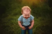 Toddler in a striped shirt and suspenders making funny face and closing his eyes while standing in tall grass.