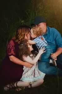 Parents kissing their toddler as his older sister reaches toward him during a candid family cuddle in a grassy field.