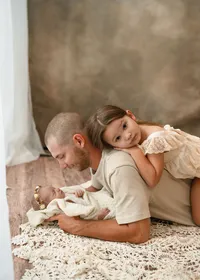 Father lying beside his newborn on a cream blanket while his older daughter rests on his back during a lifestyle newborn session.