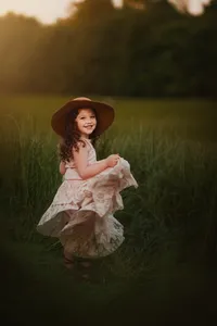 Smiling girl twirling in a flowing pink dress and brown wide-brimmed hat in tall grass during golden hour.